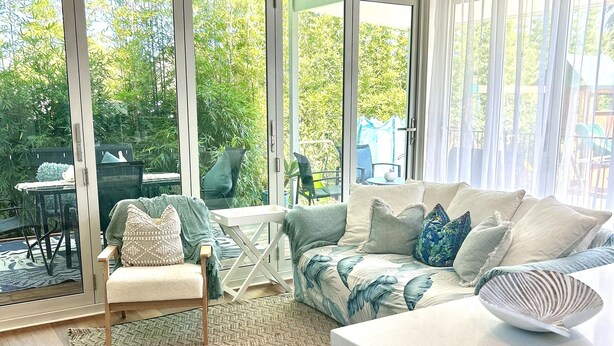 Un salón amplio con un gran ventanal corredera que da a un jardín, un sofá blanco con cojines estampados y una mesita.