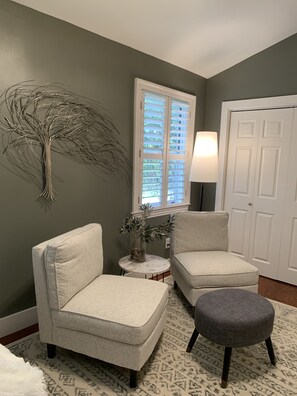 A living room with two armchairs, a small round table with a vase, a gray ottoman, a lamp, and a window with blinds.