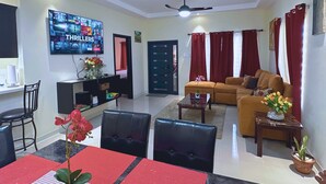A living room with a TV displaying movie posters, a sofa, a dining table with chairs, and a ceiling fan. - Pyramid Bliss - One Bedroom (Ocho Rios)