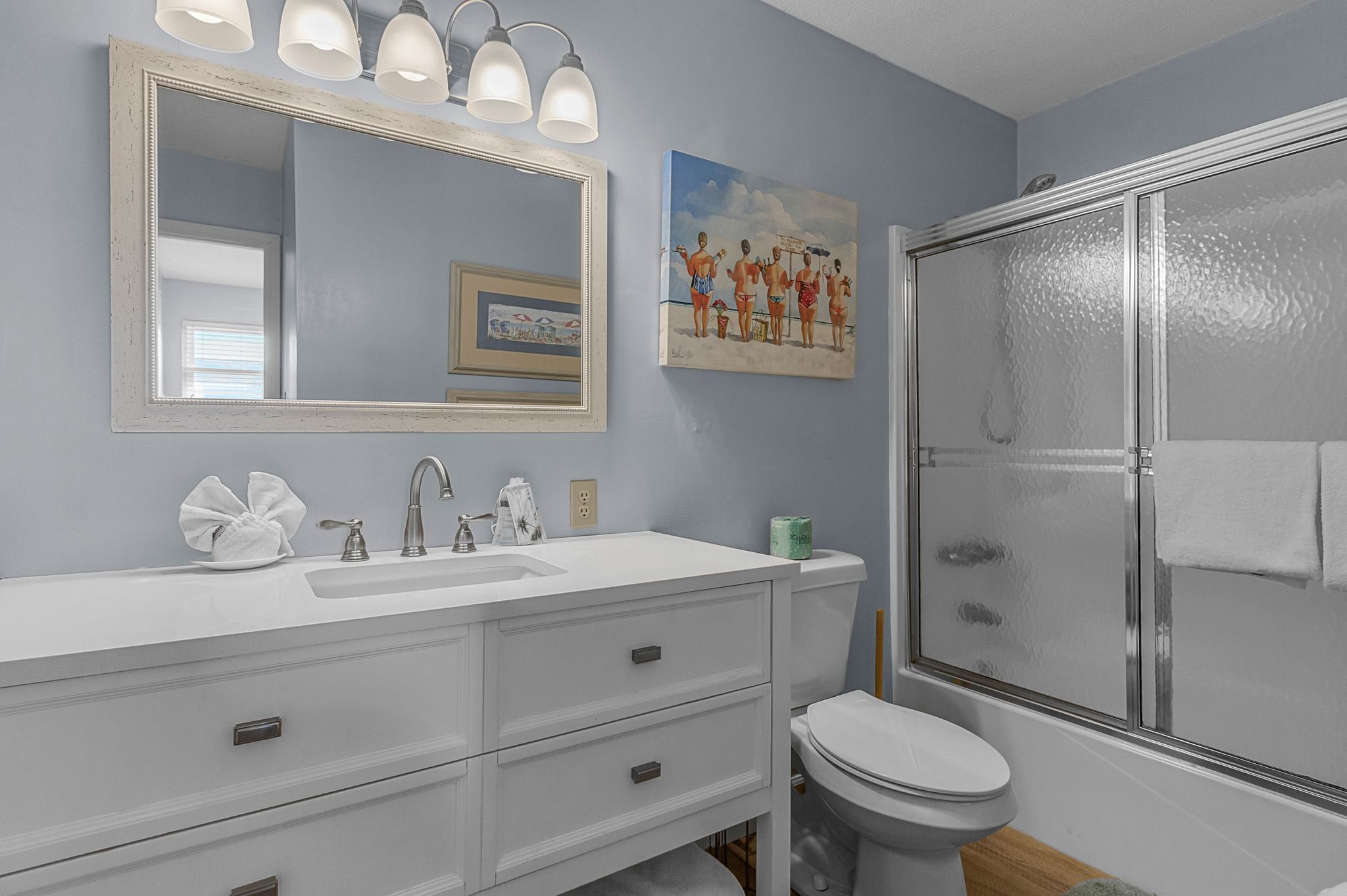 Une salle de bain avec un meuble-lavabo blanc, un grand miroir, une douche avec une porte en verre et un tableau accroché au mur.