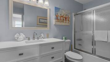 Une salle de bain avec un meuble-lavabo blanc, un grand miroir, une douche avec une porte en verre et un tableau accroché au mur.