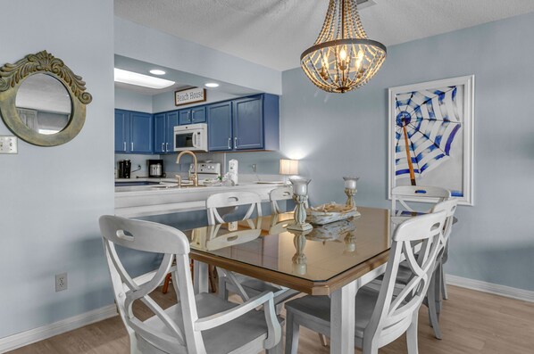 A dining area with a glass table surrounded by white chairs, a chandelier, and a kitchen in the background.