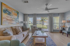 A living room with a sofa, armchairs, a coffee table, and a view of a building and palm trees through large windows.