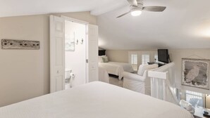 Une chambre avec un grand lit, un ventilateur de plafond et une vue sur une autre chambre équipée d’un lit et d’un téléviseur.