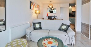 A living room with a sofa, a coffee table, a lamp, a painting, and a kitchen in the background.