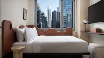 A modern hotel room with a large bed, a side table, a small round ottoman, a desk with a lamp, and a city view through a large window.
