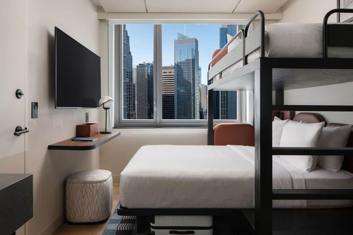 A compact hotel room with bunk beds, a flat-screen TV, a small desk with a lamp, and a window offering a city view.