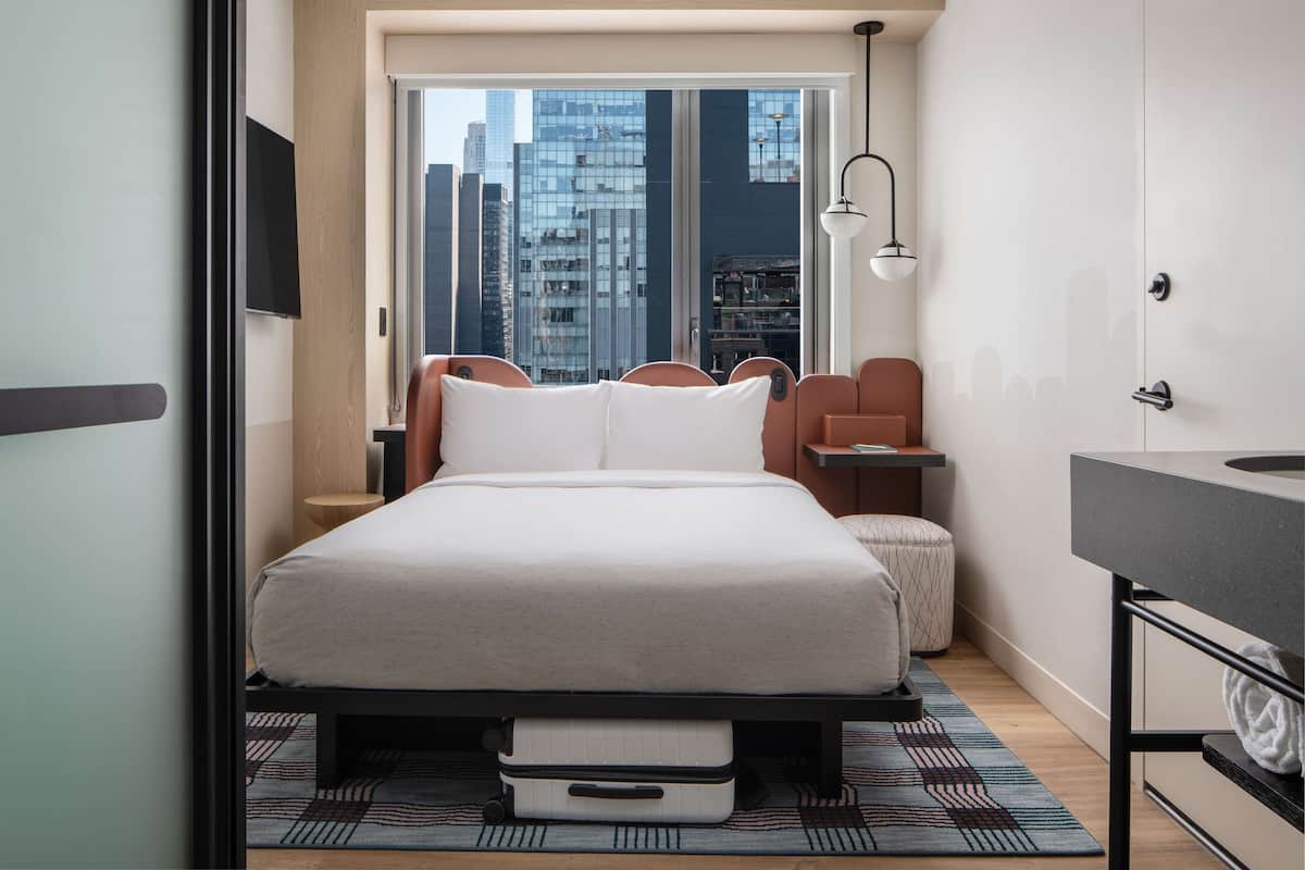 A modern hotel room with a large bed, a desk, a chair, a small table, a bench, a window with a city view, and a bathroom with a mirror and sink.