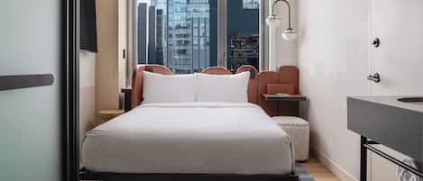 A modern hotel room with a large bed, a desk, a chair, a small table, a bench, a window with a city view, and a bathroom with a mirror and sink.