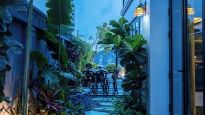A narrow pathway with stepping stones leading to a seating area with chairs and tables, surrounded by lush tropical plants and a building with large windows. - Indochine‑Style Private Pool Villa Full Bay View |5 BR | Karaoke • Billiards (Thành phố Hạ Long)
