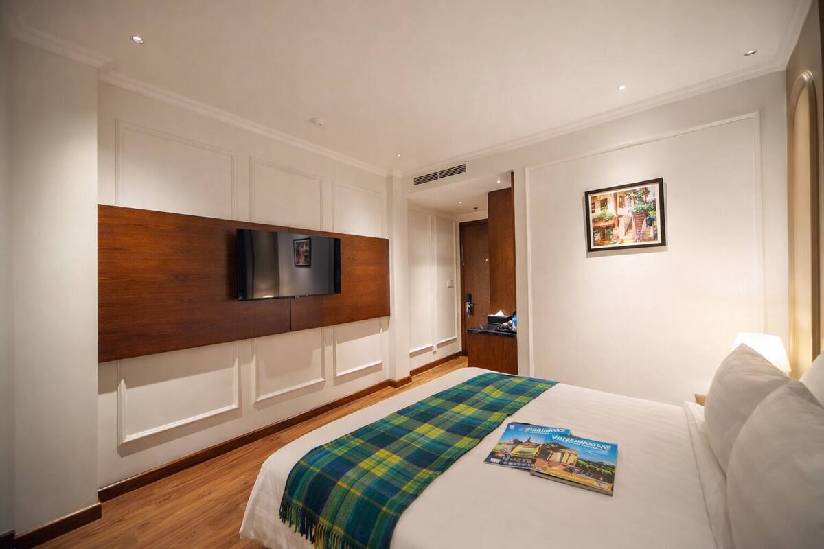 A hotel room with a large bed, a mounted TV, a wooden headboard, a door leading to a bathroom, and a framed picture on the wall.
