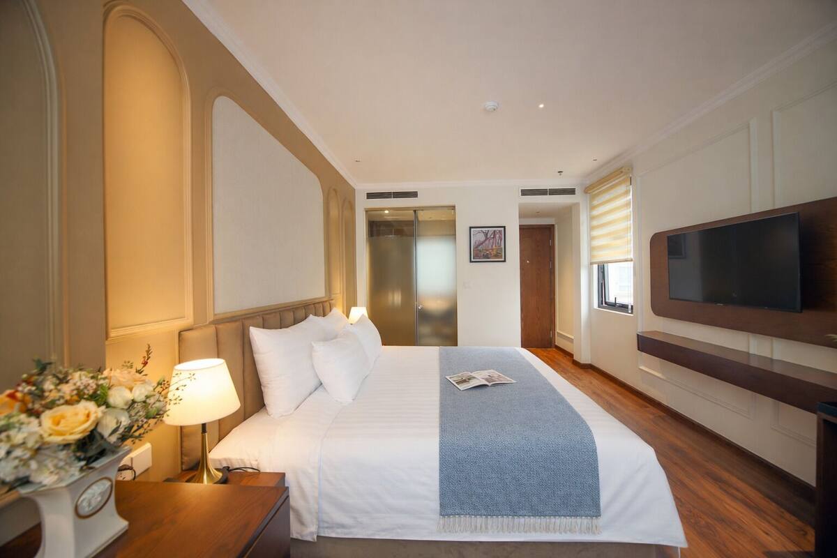 A hotel room with a large bed, a TV mounted on the wall, a bedside table with a lamp, and a door leading to a bathroom.