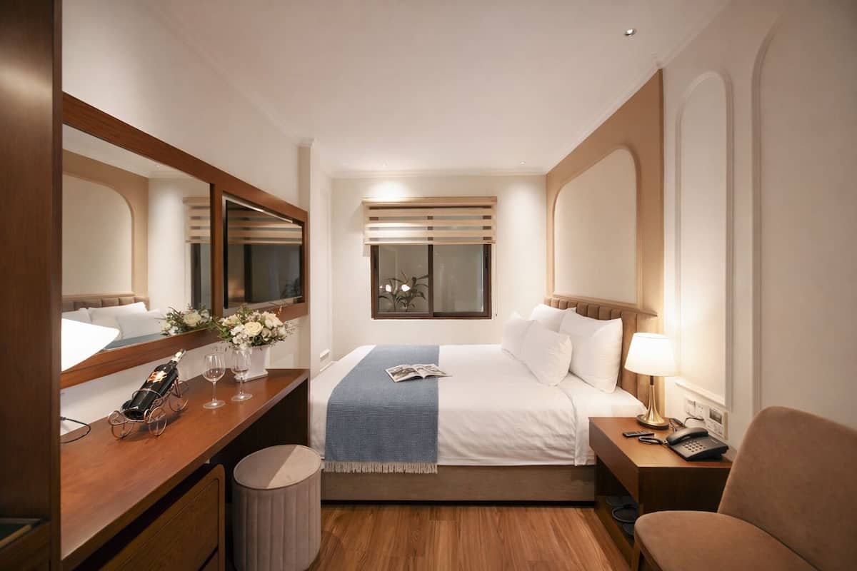 A hotel room with a bed, bedside table, lamp, chair, and a desk with a telephone.