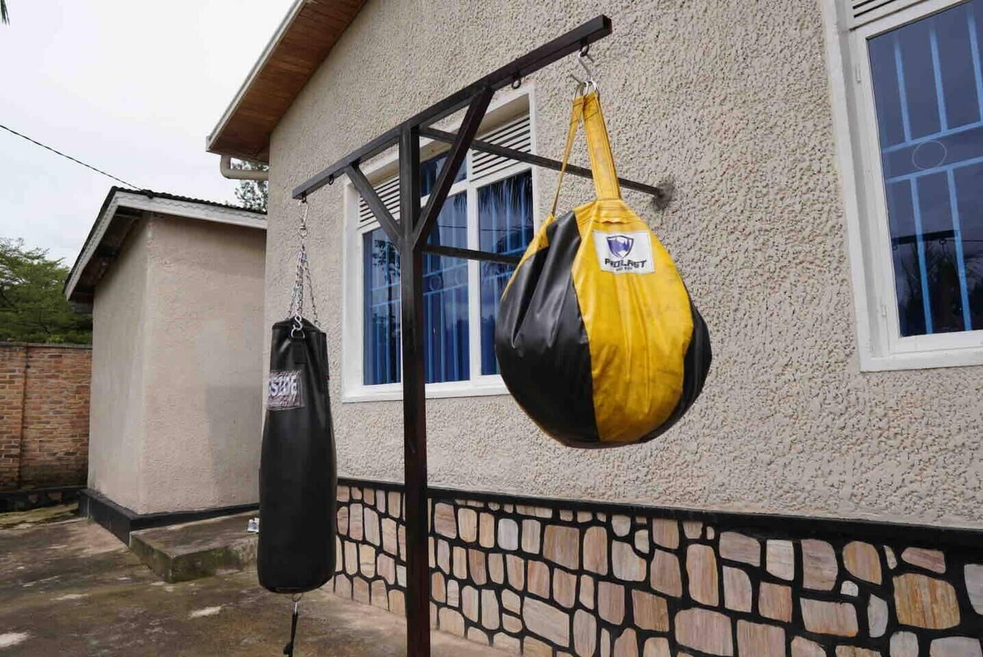 Deux sacs de frappe sont suspendus à une structure métallique devant une maison dont la fondation est constituée d’un mur en pierre et qui possède une fenêtre ornée de rideaux bleus.