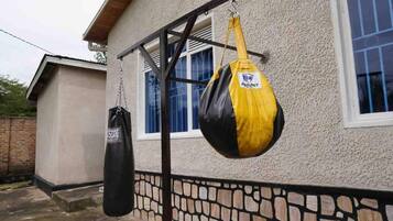 Deux sacs de frappe sont suspendus à une structure métallique devant une maison dont la fondation est constituée d’un mur en pierre et qui possède une fenêtre ornée de rideaux bleus.