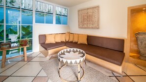 A modern living room with a brown sectional sofa, a round woven coffee table, and a large window with a view of greenery. - Villa Panchajanya (Seminyak)