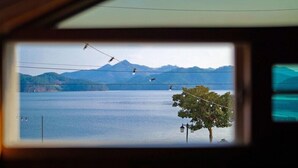 Vista a través de una ventana que muestra un lago con montañas al fondo y un árbol adornado con luces colgantes.