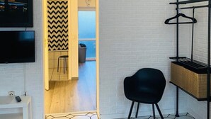 Una habitación con pared de ladrillo blanco, un televisor montado en ella, una silla negra, suelo de madera y una puerta que da a otra habitación.