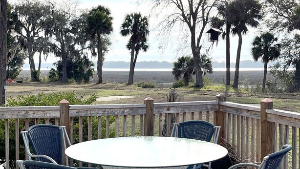 A covered patio with a round table and four chairs, overlooking a natural landscape with trees and a distant view of a body of water.