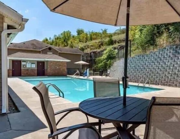 A swimming pool with lounge chairs and umbrellas, a brick wall, and a building with a door and windows.
