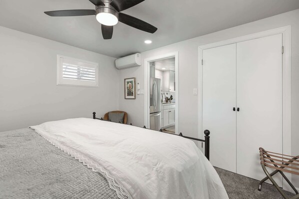 A bedroom with a bed, a ceiling fan, a window with blinds, a white door, and a bathroom visible through an open door.