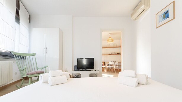 A modern hotel room with a bed, a television, a green rocking chair, and a dining area visible through an open door. - Via Marcona 17 Apartment (Milano)