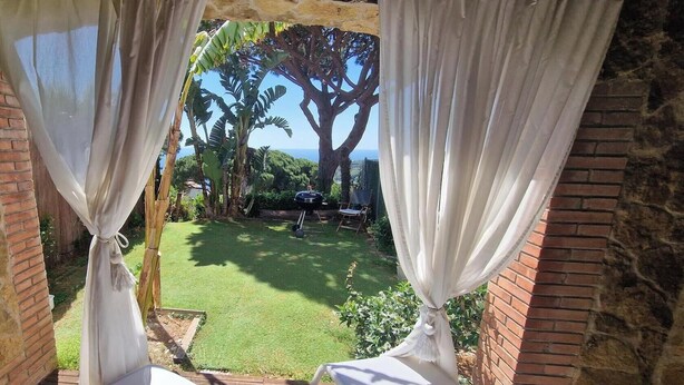 A view through a window with white curtains, a grassy yard, a tree, and a brick wall. - Mediterranean house with SEA VIEWS, garden and terrace. (Sant Feliu de Guíxols)