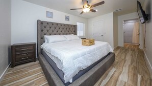 A bedroom with a tufted headboard, a bed with white linens, a bedside table with drawers, a ceiling fan, and a television mounted on the wall. - Casa flower centrally located  (Tucson)