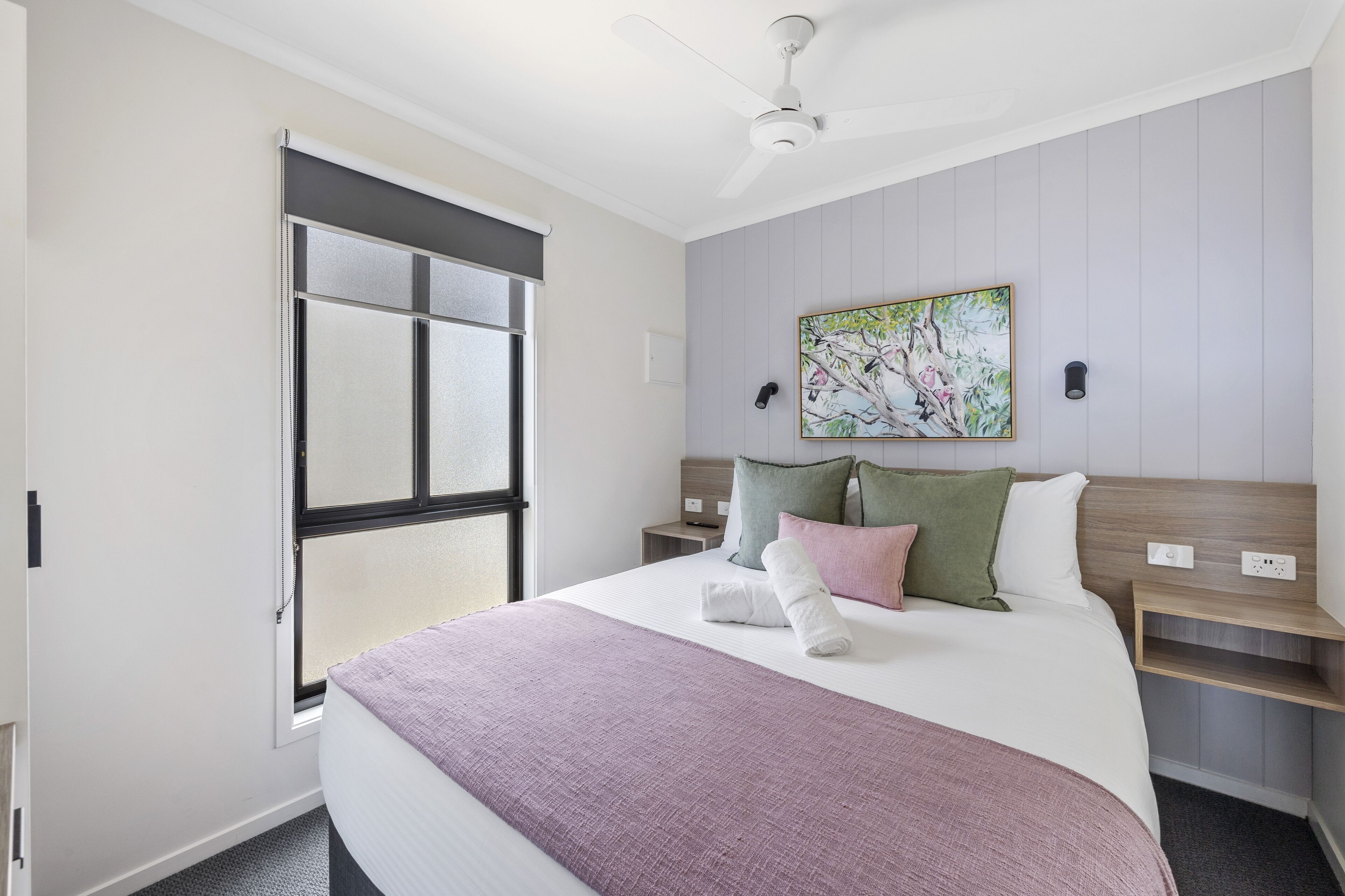 A hotel room with a bed, a nightstand, a wall-mounted shelf, a ceiling fan, and a window with blinds.