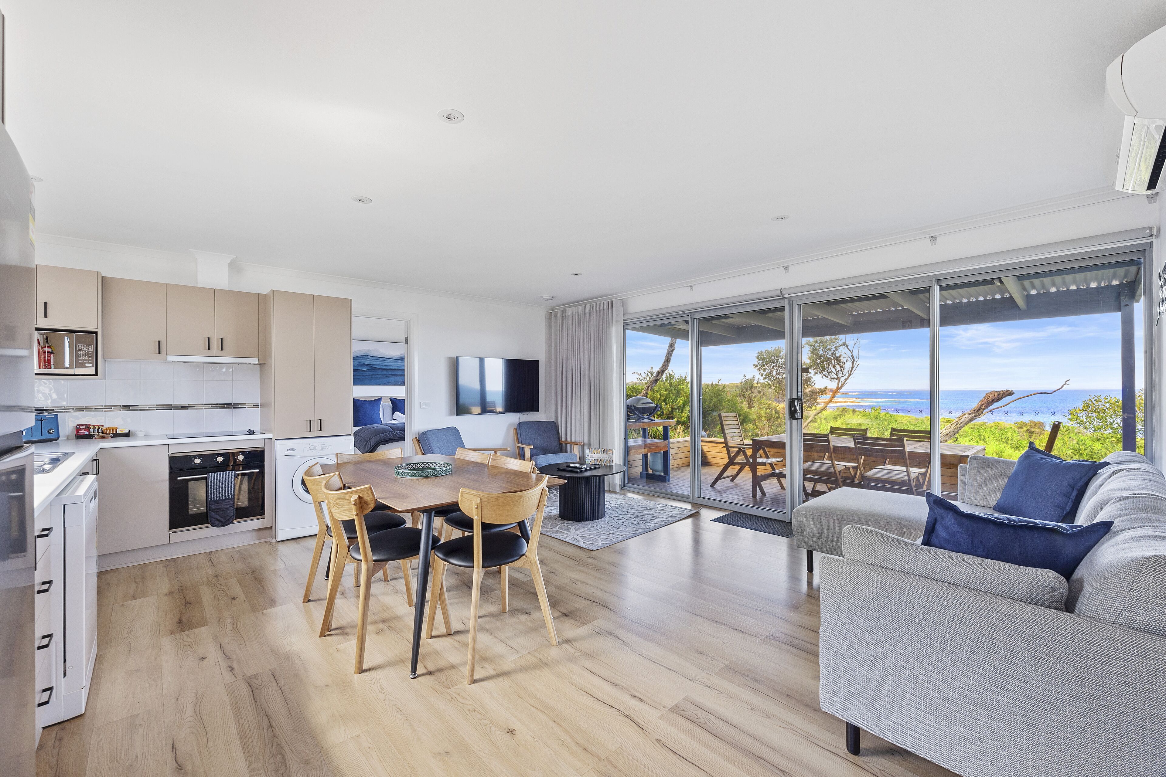 Una cocina moderna con armarios blancos, un comedor con mesa y sillas de madera, un sofá gris y un amplio ventanal corredera que da a un balcón con vistas al mar.