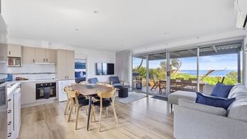 Una cocina moderna con armarios blancos, un comedor con mesa y sillas de madera, un sofá gris y un amplio ventanal corredera que da a un balcón con vistas al mar.