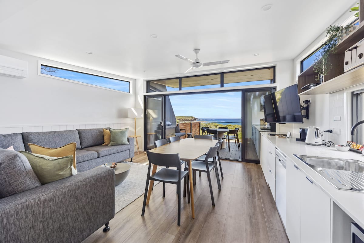A modern kitchen with a large island, a dining area with a view, and a cozy seating area with a sofa and cushions.