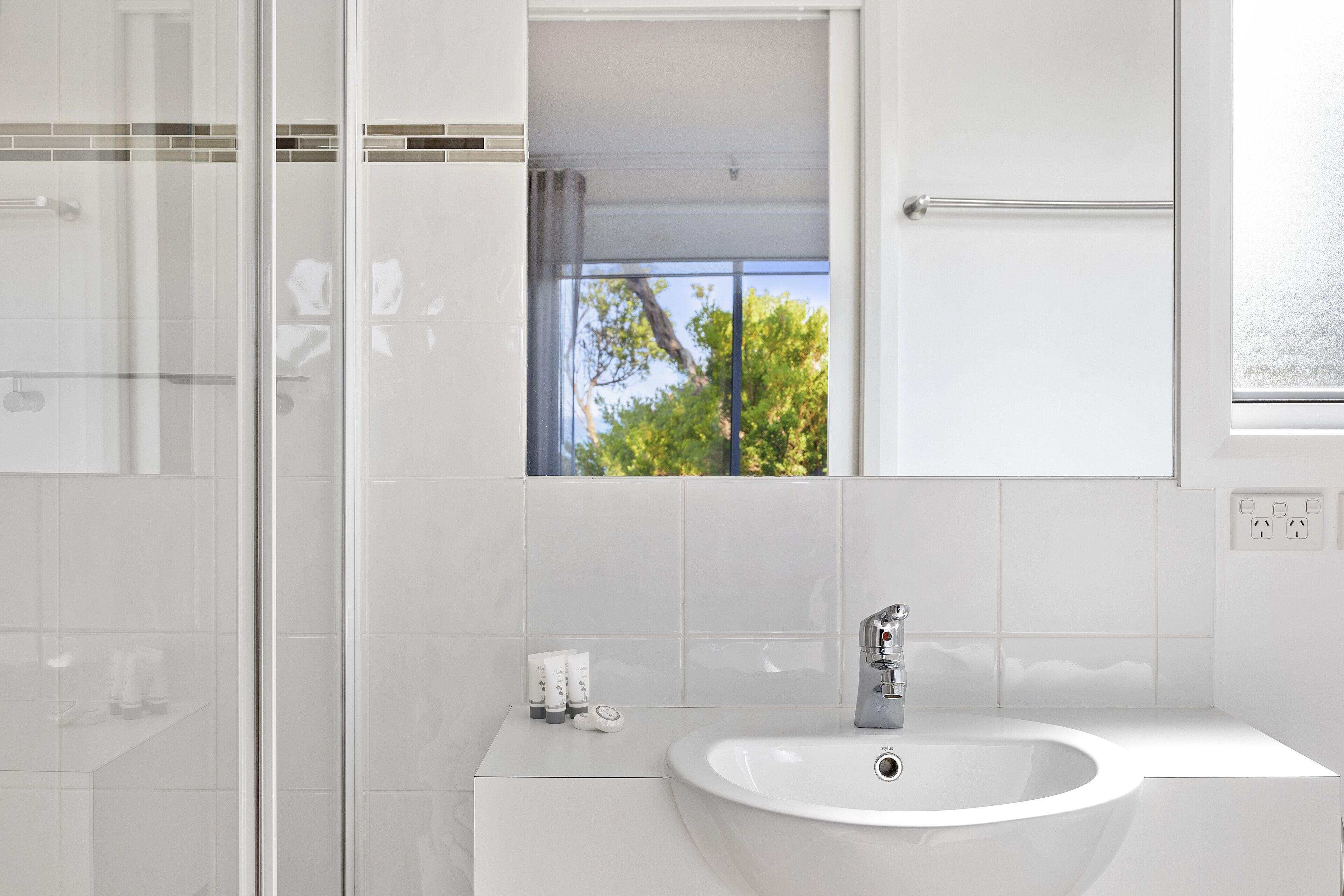 A modern bathroom with a glass shower enclosure, a white sink, and a window with a view of trees.