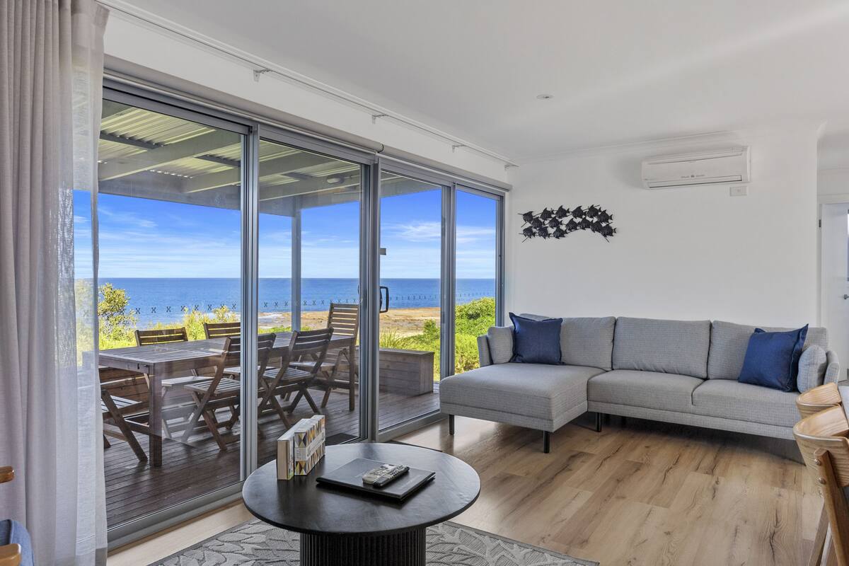 A modern living room with a grey sofa, a round coffee table, and a view of the sea through large sliding glass doors.
