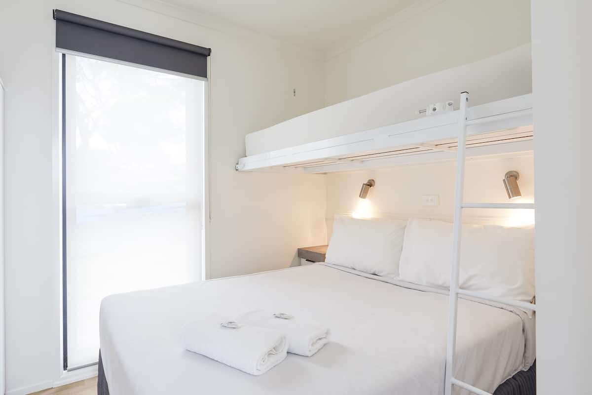 A bunk bed with white bedding, a nightstand, a lamp, and a window with blinds.