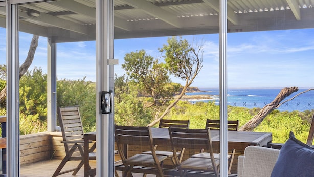A modern room with a large sliding glass door leading to an outdoor area with a view of the ocean. There is a sofa and a dining table with chairs inside the room. - Ingenia Holidays Cape Paterson (Cape Paterson)