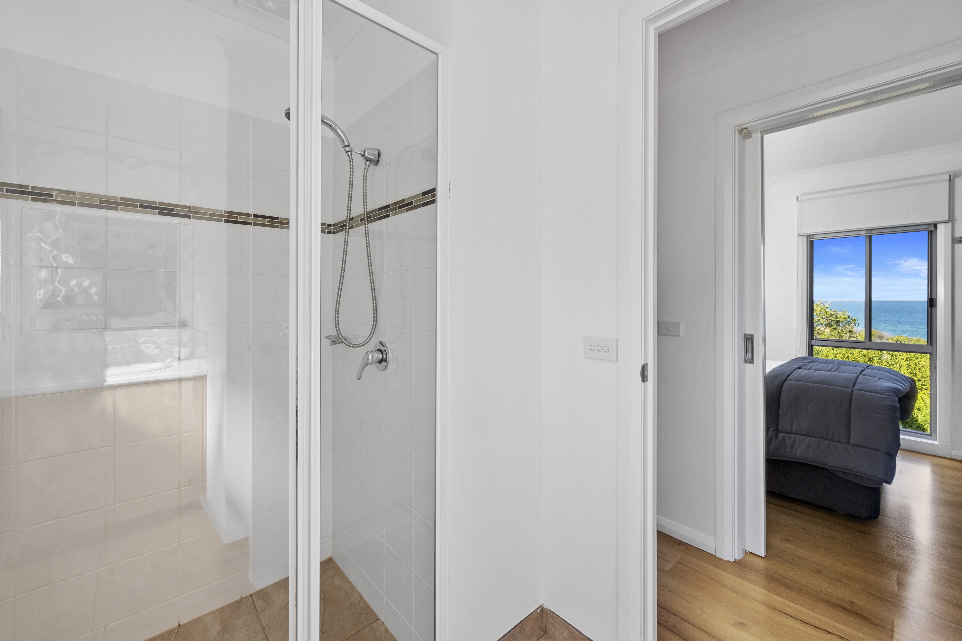 A modern bathroom with a glass shower enclosure, a visible towel rack, and a view of the ocean through an open door.