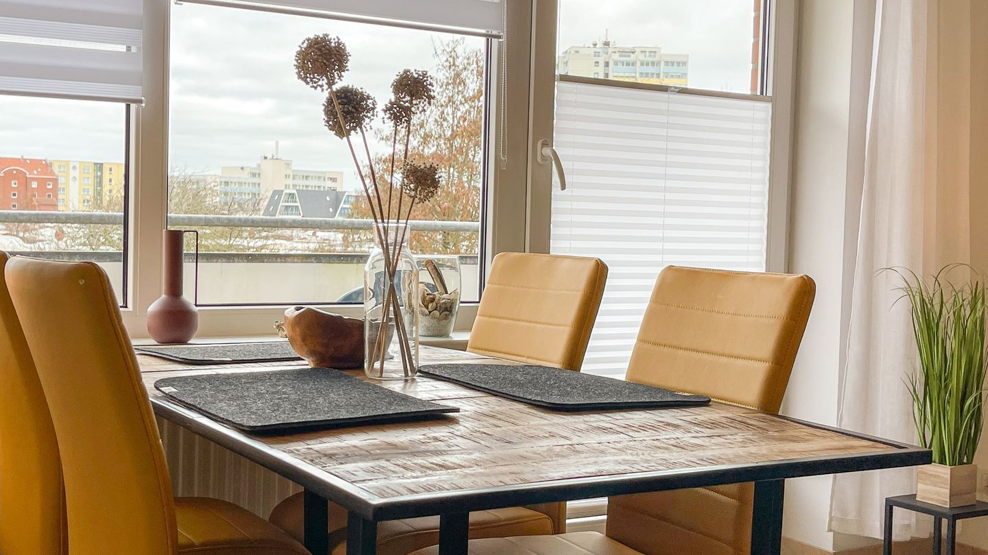 Ein Essbereich mit einem Tisch für vier Personen, je zwei Stühle an jeder Seite, einer Vase mit getrockneten Blumen und Blick auf Gebäude durch große Fenster.