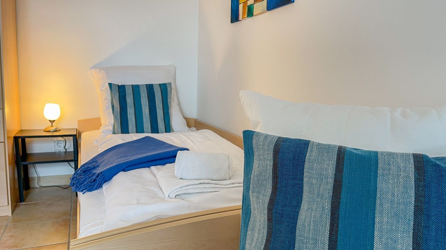 Ein ordentlich bezogenes Bett mit blau-weiß gestreiften Kissen, einer blauen Decke und einem weißen Handtuch. Ein kleiner Nachttisch mit Lampe und Steckdose ist ebenfalls vorhanden.