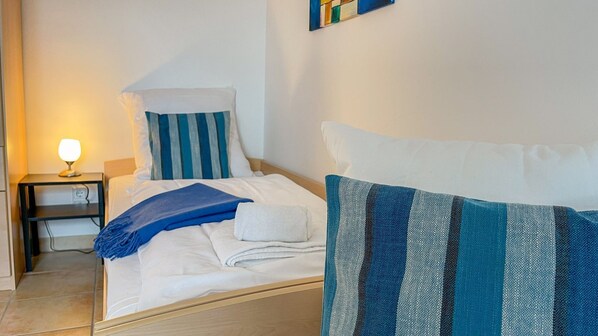 Un lit bien fait, avec des oreillers à rayures blanches et bleues, une couverture bleue et une serviette blanche. On y trouve une petite table de chevet avec une lampe et une prise de courant.