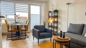 Ein modernes Wohnzimmer mit einem grauen Sofa, einem runden Holztisch, einem grauen Sessel und einem Essbereich mit Tisch und Stühlen.