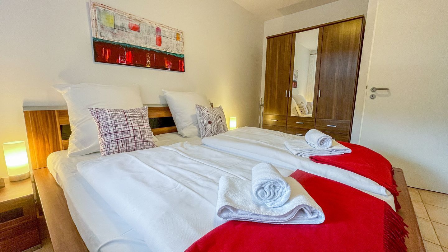 Ein ordentlich bezogenes Bett mit weißen Leinentüchern und einer roten Decke, zwei zusammengerollte Handtücher und ein Holzschrank in einem Hotelzimmer.