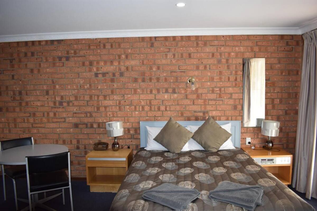 A hotel room with a brick wall, a bed with a patterned bedspread, two bedside tables with lamps, a small table with a chair, and a window with curtains.