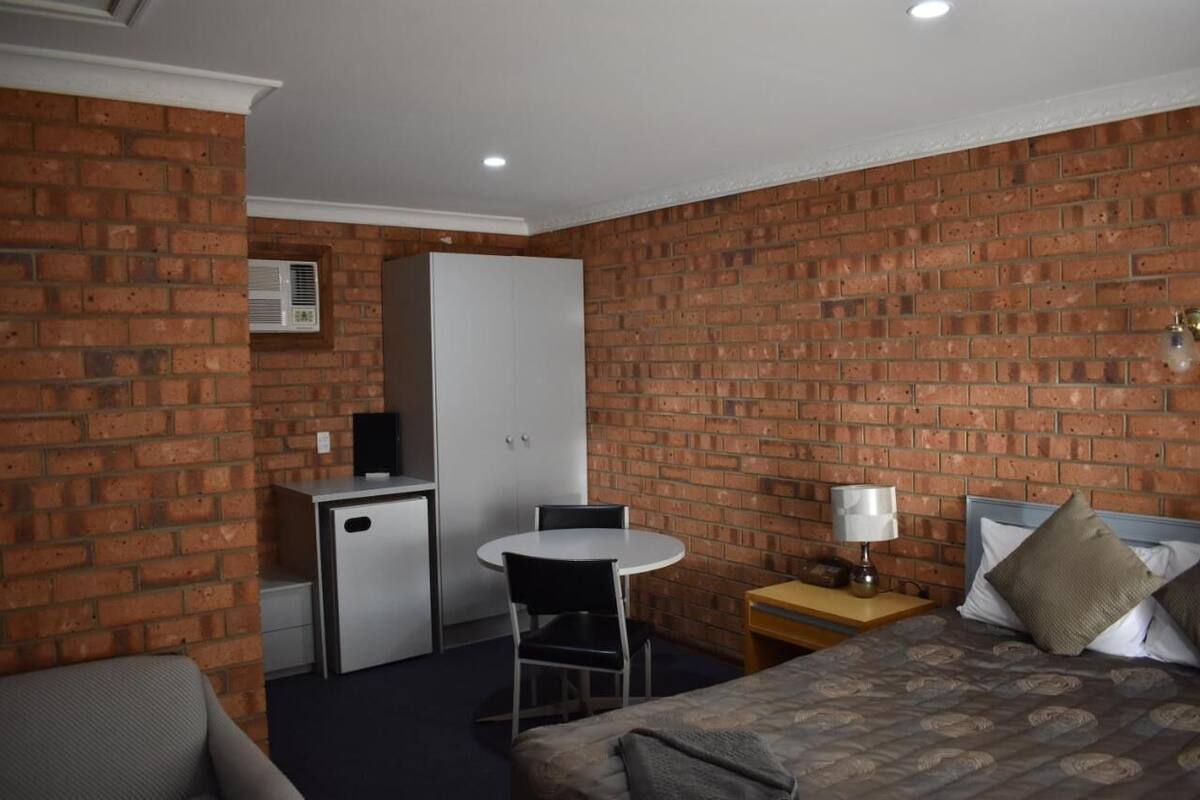 A hotel room with a brick wall, a bed with pillows, a desk with a chair, a small kitchenette, and a window.