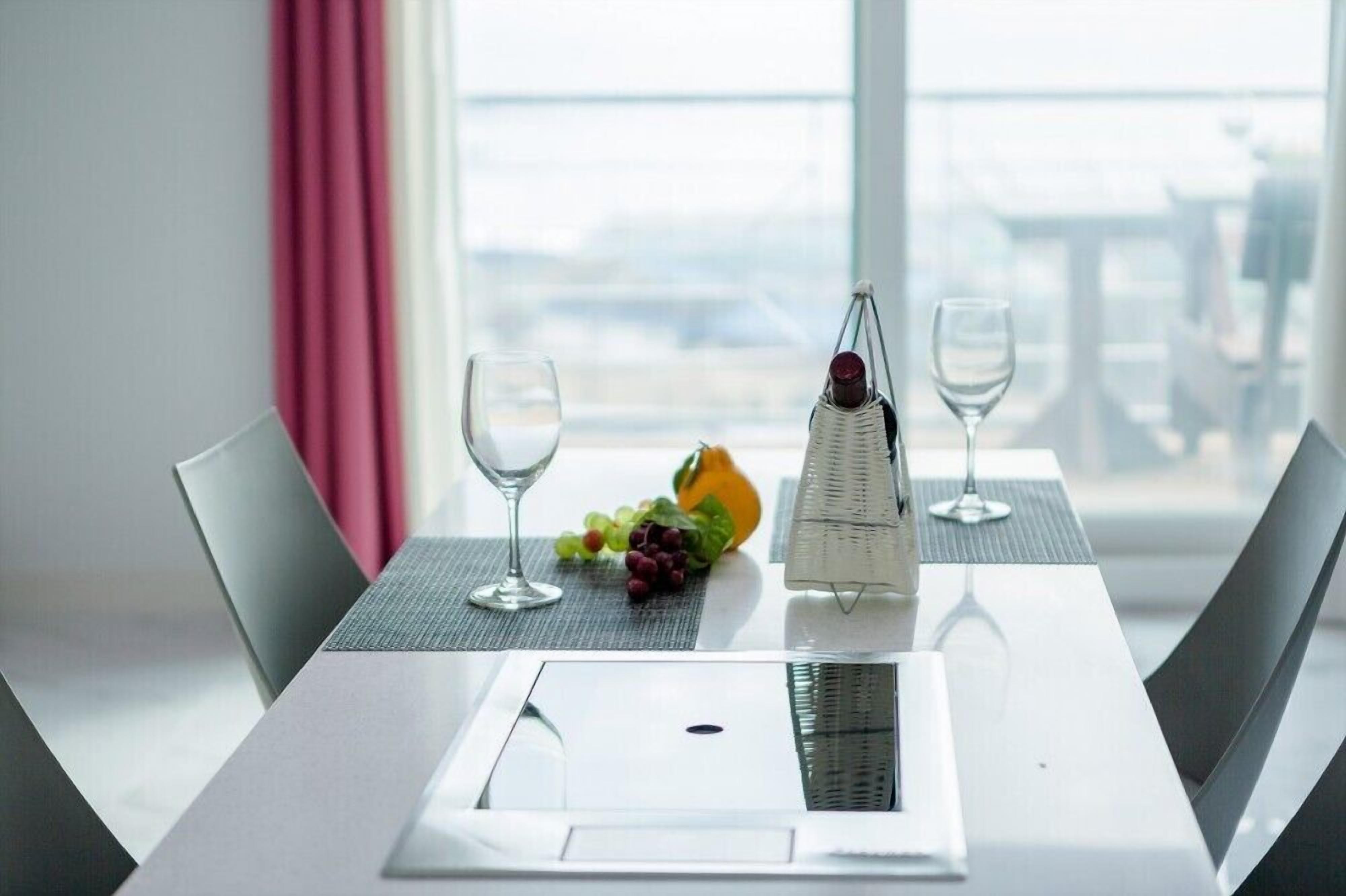 A hotel room with a dining table set for two, a bottle of wine, and a fruit bowl with grapes and a melon.