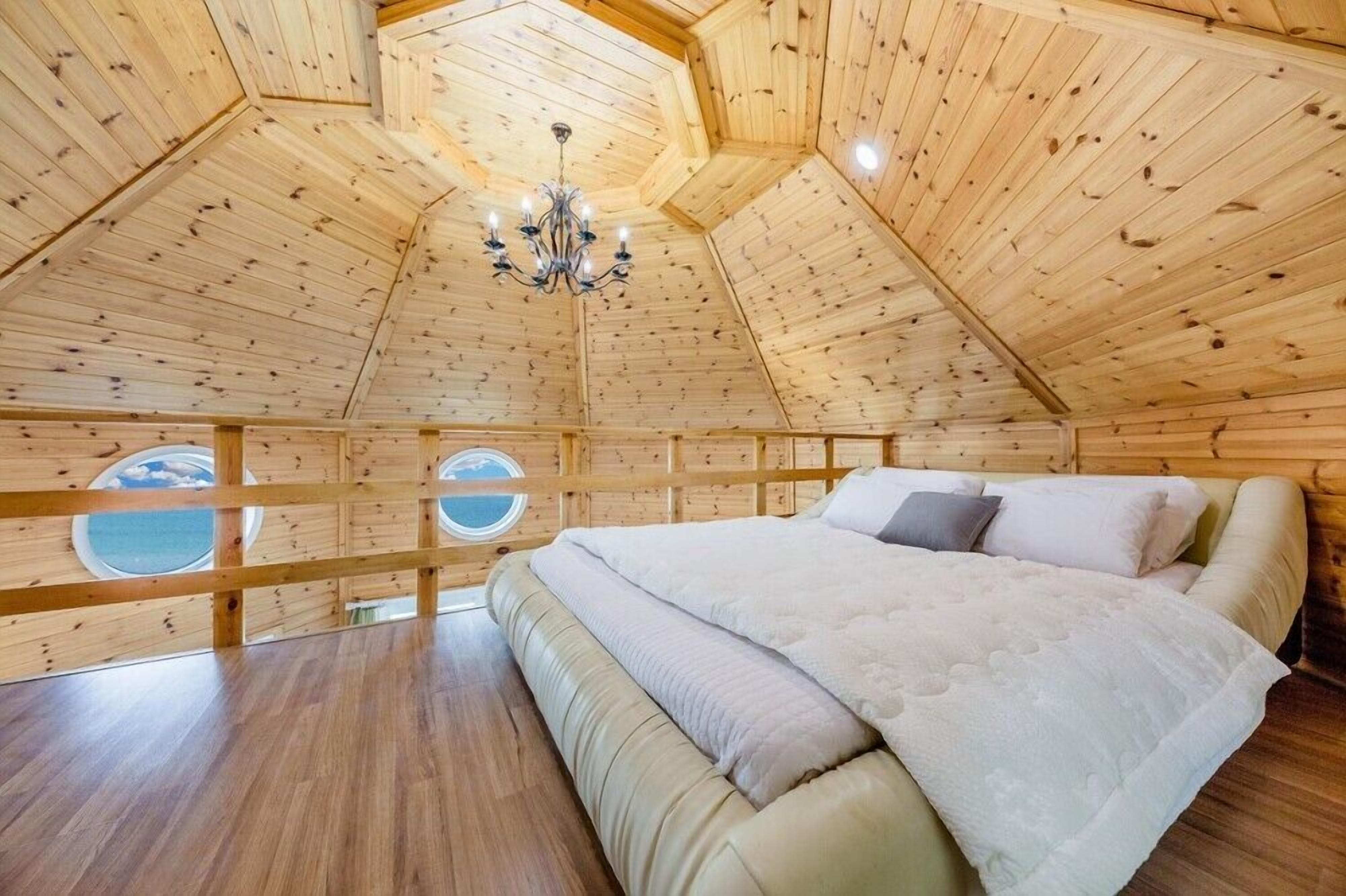 Un intérieur chaleureux, avec des murs en bois et un plafond haut, un lit recouvert d’une couette blanche et une fenêtre ronde.