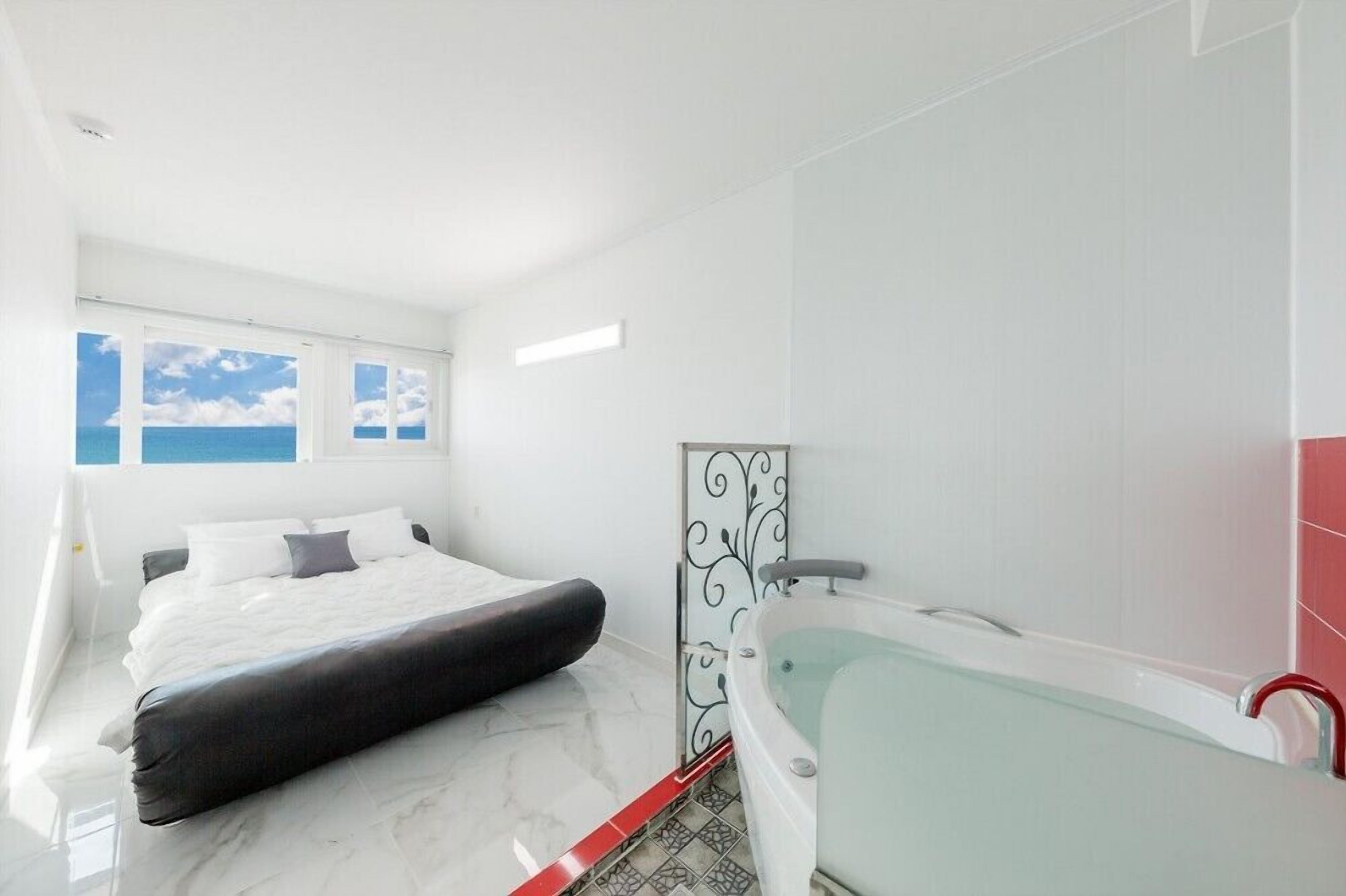 A modern bedroom with a large bed, a glass-enclosed bathtub, and a view of the sky through the windows.