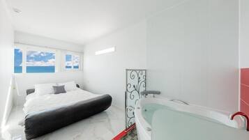 A modern bedroom with a large bed, a glass-enclosed bathtub, and a view of the sky through the windows.