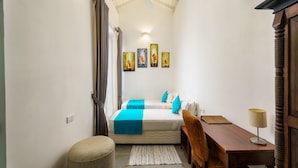 Ein kleiner, gut beleuchteter Raum mit einem Bett, einem Schreibtisch und einem Sessel. An der Wand hängen drei Bilder und eine Deckenventilator ist angebracht.