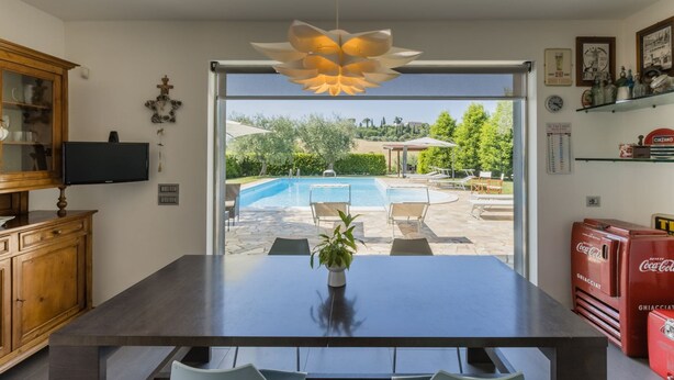 A modern kitchen with a large dining table, a TV, and a view of an outdoor swimming pool.